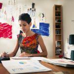 Money and financial planning, young hispanic self-employed woman checking bills and doing budget with calculator, computer and papers in fashion design studio