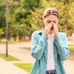A young woman walking through a park in spring, rubbing her nose and looking uncomfortable because of allergies.