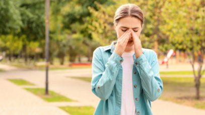 A young woman walking through a park in spring, rubbing her nose and looking uncomfortable because of allergies.