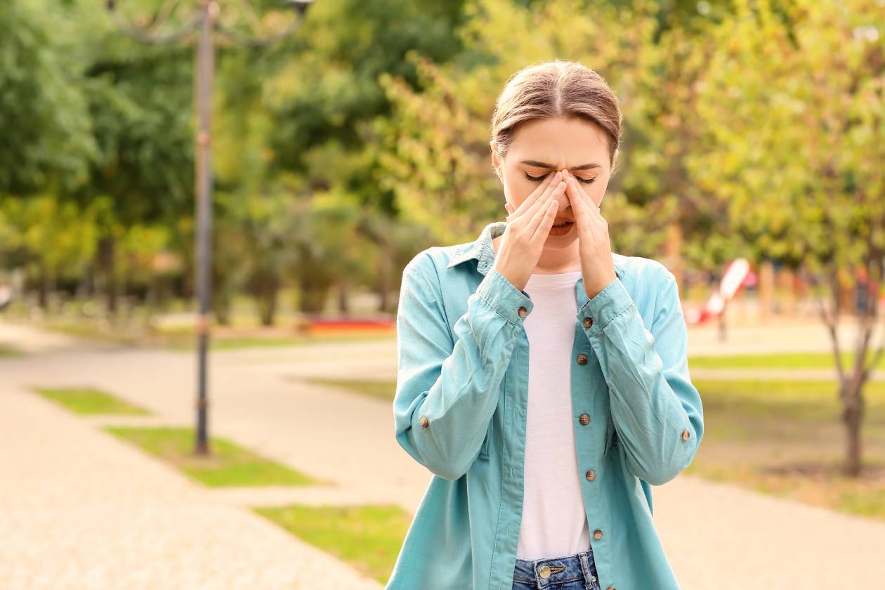 A young woman walking through a park in spring, rubbing her nose and looking uncomfortable because of allergies.