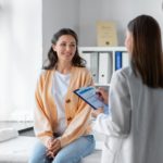 A dark haired woman in an orange cardigan having a consultation with a doctor during her hospital visit.