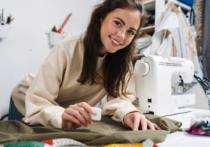 A young brunette woman smiling at the camera as she uses chalk to mark a piece of fabric for a sewing project.