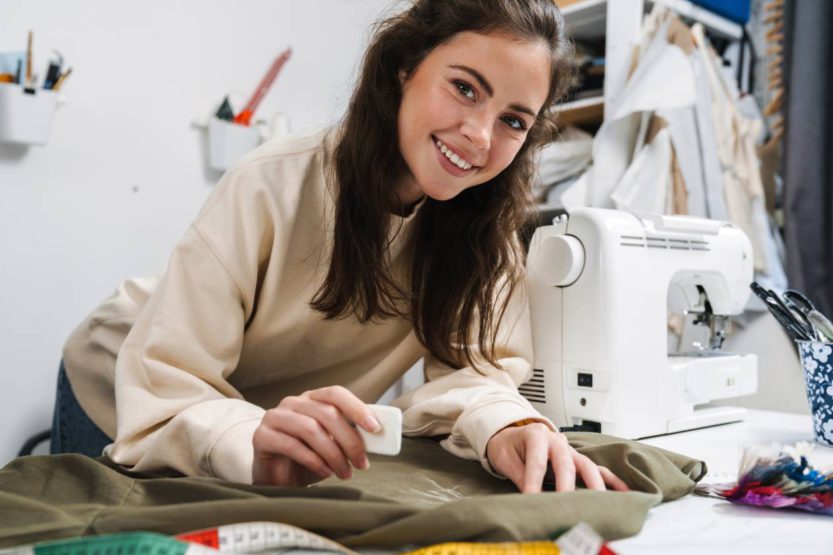 A young brunette woman smiling at the camera as she uses chalk to mark a piece of fabric for a sewing project.