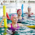 Three women wearing bathing suits, swim caps, and goggles in a pool. They are holding pool noodles and smiling.