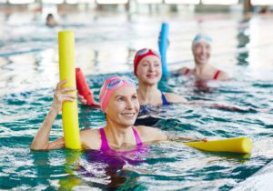 Three women wearing bathing suits, swim caps, and goggles in a pool. They are holding pool noodles and smiling.