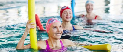 Three women wearing bathing suits, swim caps, and goggles in a pool. They are holding pool noodles and smiling.