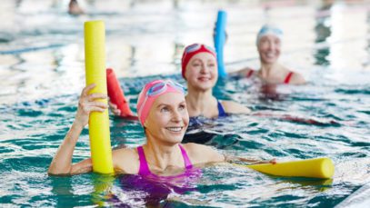 Three women wearing bathing suits, swim caps, and goggles in a pool. They are holding pool noodles and smiling.