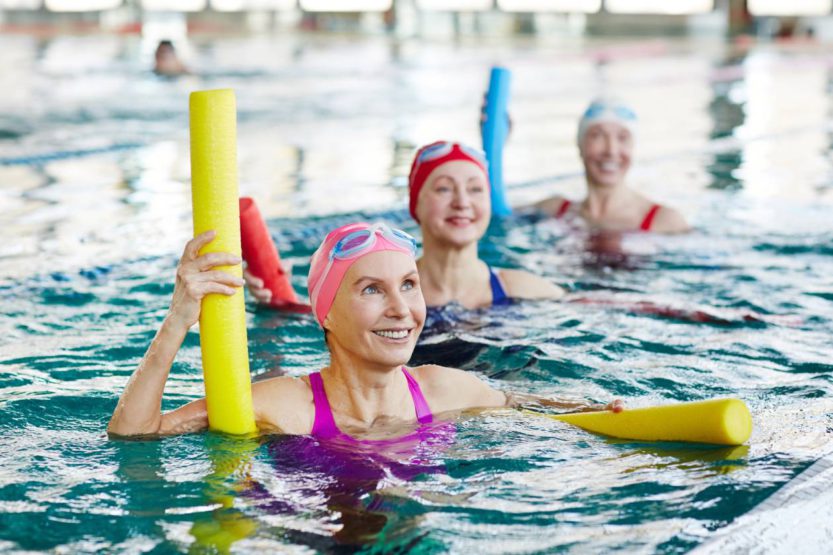 Three women wearing bathing suits, swim caps, and goggles in a pool. They are holding pool noodles and smiling.