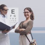 A young woman is wearing glasses and smiling while her optometrist, another woman in glasses and a lab coat, speaks to her.