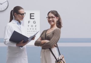 A young woman is wearing glasses and smiling while her optometrist, another woman in glasses and a lab coat, speaks to her.