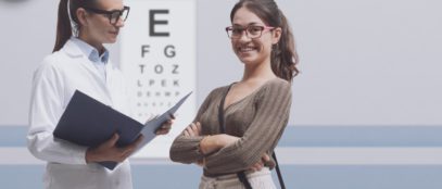 A young woman is wearing glasses and smiling while her optometrist, another woman in glasses and a lab coat, speaks to her.