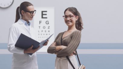 A young woman is wearing glasses and smiling while her optometrist, another woman in glasses and a lab coat, speaks to her.