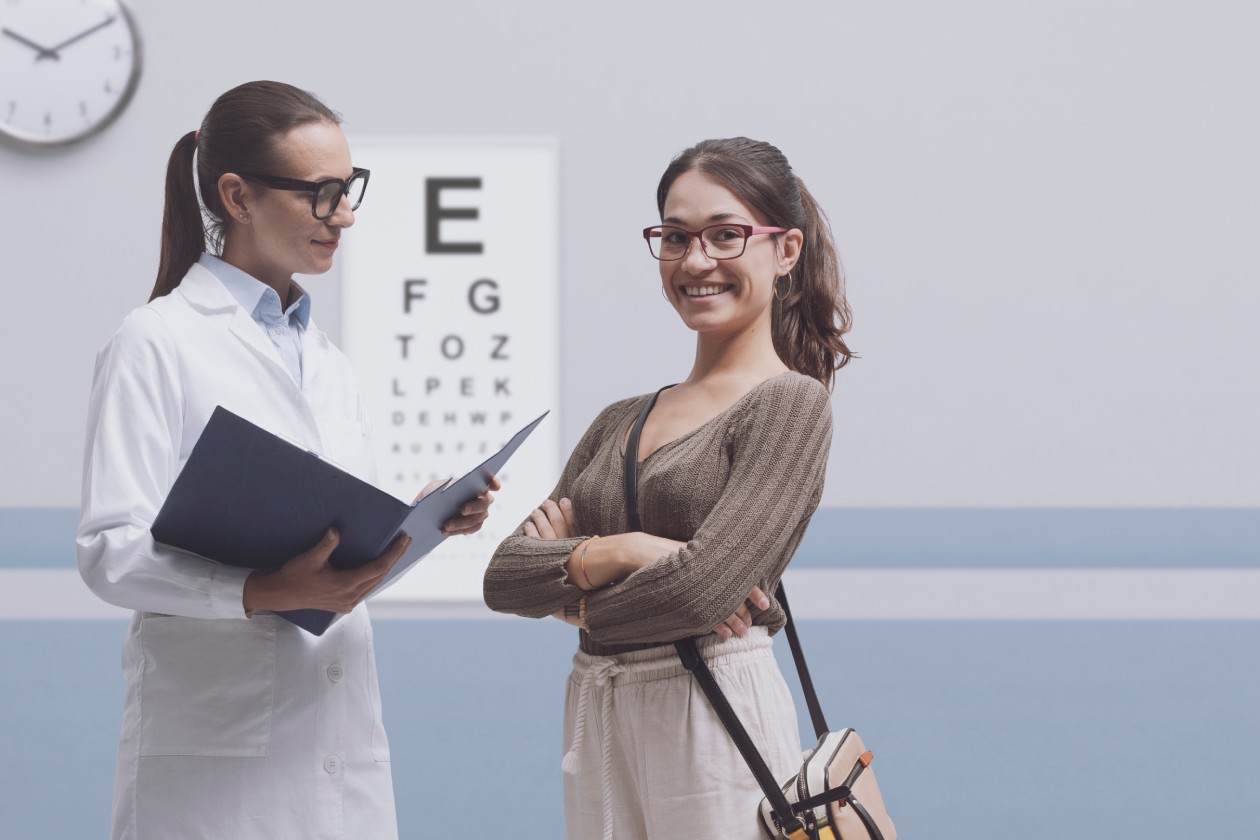 A young woman is wearing glasses and smiling while her optometrist, another woman in glasses and a lab coat, speaks to her.
