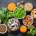 On a table full of vegetables, ten bowls contain various legume, nut, and bean plant-based protein sources.