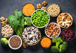 On a table full of vegetables, ten bowls contain various legume, nut, and bean plant-based protein sources.