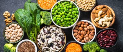 On a table full of vegetables, ten bowls contain various legume, nut, and bean plant-based protein sources.