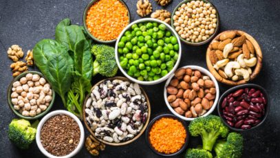 On a table full of vegetables, ten bowls contain various legume, nut, and bean plant-based protein sources.