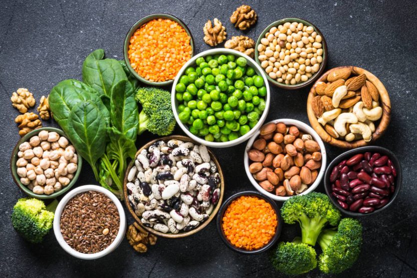 On a table full of vegetables, ten bowls contain various legume, nut, and bean plant-based protein sources.
