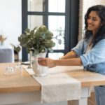 A woman wearing a denim outfit who’s embracing simplicity at home and decorating her kitchen table with a white vase and plants.