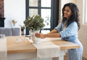 A woman wearing a denim outfit who’s embracing simplicity at home and decorating her kitchen table with a white vase and plants.