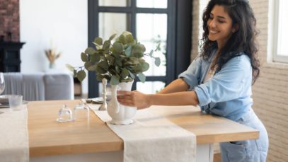 A woman wearing a denim outfit who’s embracing simplicity at home and decorating her kitchen table with a white vase and plants.