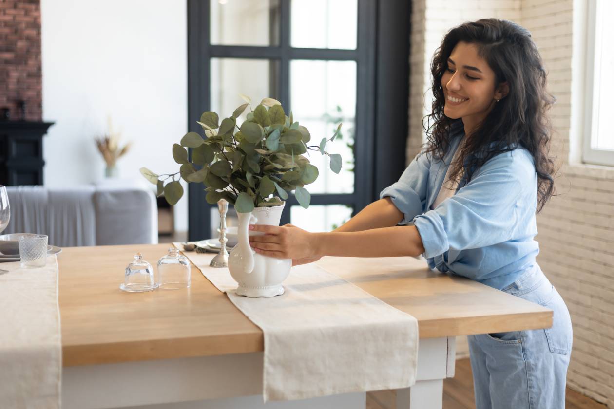 A woman wearing a denim outfit who’s embracing simplicity at home and decorating her kitchen table with a white vase and plants.