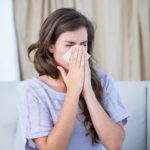 A sick woman sitting on a couch in her living room wearing a knit, purple shirt blows her nose into a tissue.