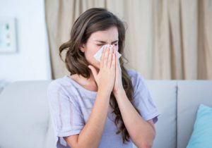 A sick woman sitting on a couch in her living room wearing a knit, purple shirt blows her nose into a tissue.