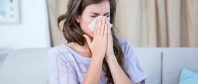 A sick woman sitting on a couch in her living room wearing a knit, purple shirt blows her nose into a tissue.