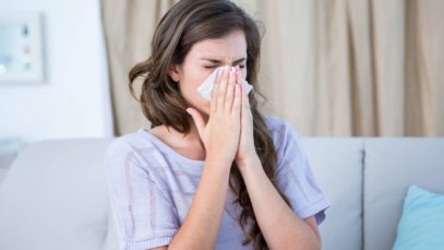 A sick woman sitting on a couch in her living room wearing a knit, purple shirt blows her nose into a tissue.