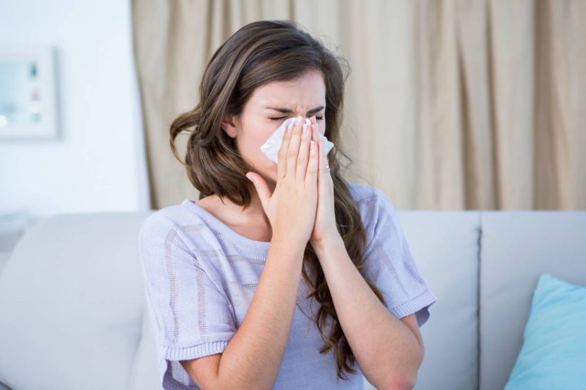 A sick woman sitting on a couch in her living room wearing a knit, purple shirt blows her nose into a tissue.
