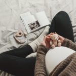 A close-up of a mother's pregnant belly. She is dressed in comfortable clothing and holds a small teddy bear over her belly.