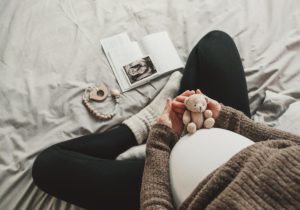 A close-up of a mother's pregnant belly. She is dressed in comfortable clothing and holds a small teddy bear over her belly.