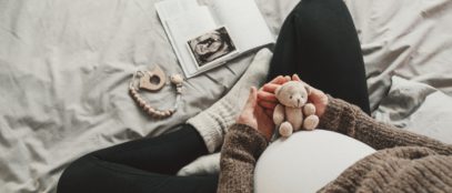 A close-up of a mother's pregnant belly. She is dressed in comfortable clothing and holds a small teddy bear over her belly.
