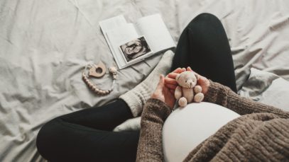 A close-up of a mother's pregnant belly. She is dressed in comfortable clothing and holds a small teddy bear over her belly.
