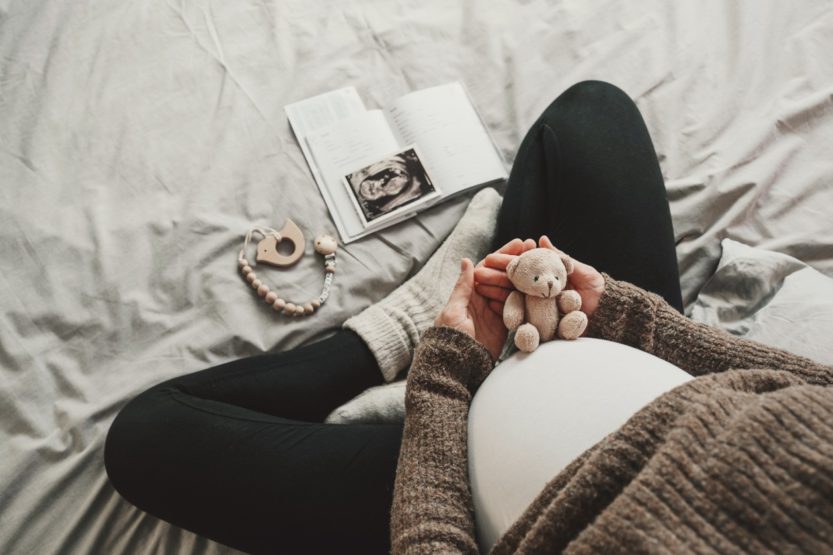 A close-up of a mother's pregnant belly. She is dressed in comfortable clothing and holds a small teddy bear over her belly.